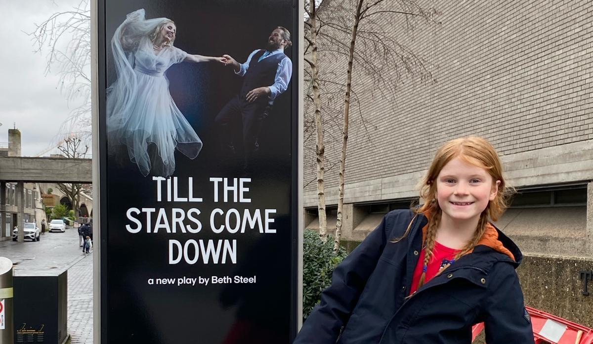 Maggie Livermore next to an advert for 'Till the Stars Come Down' at the National Theatre