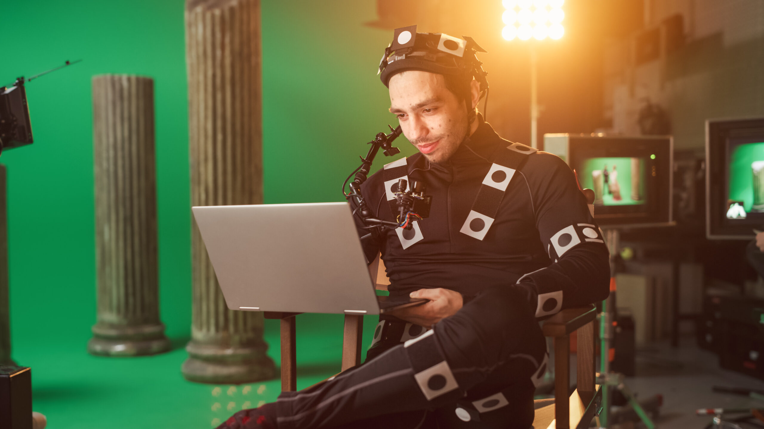 An actor dressed in a mo-cap suit looking at a laptop on set