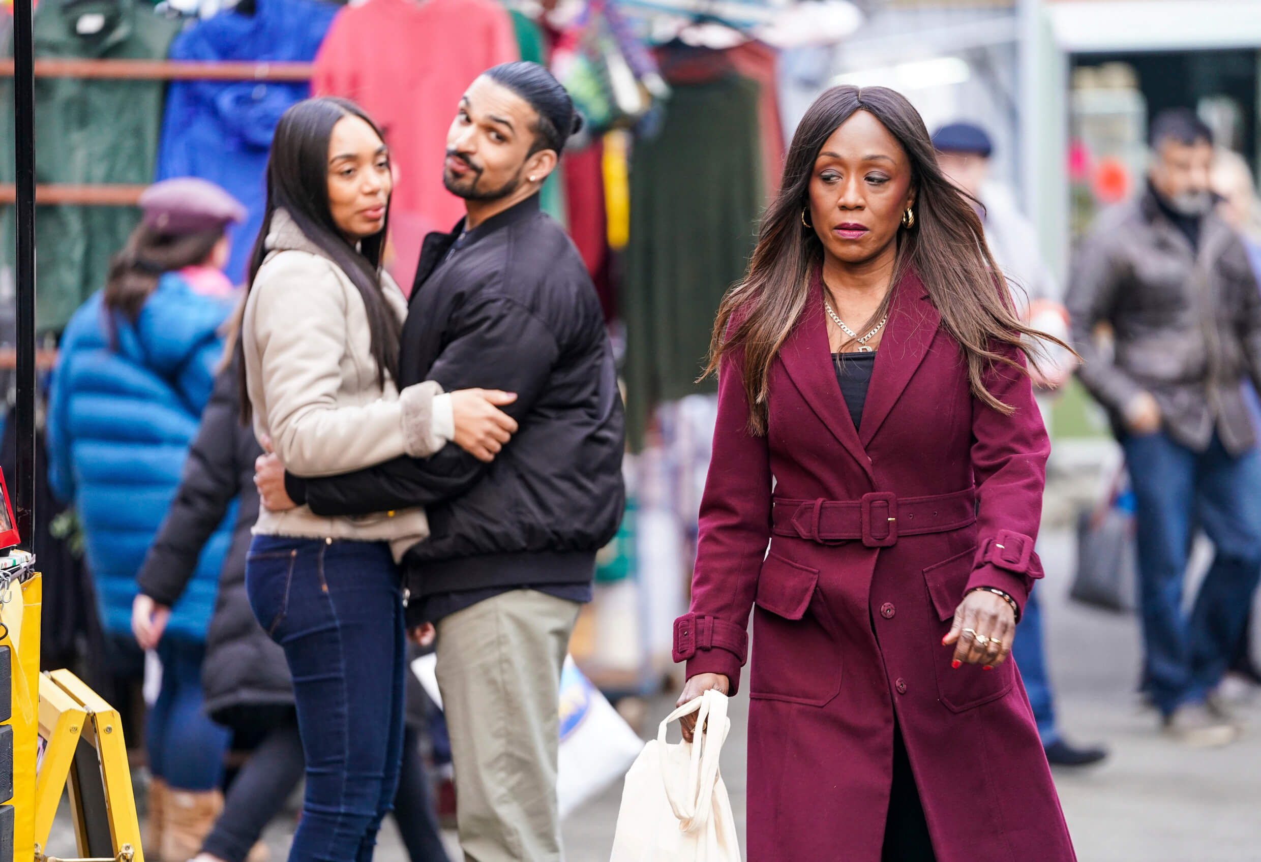 Denise in 'EastEnders' walking through the market while Chelsea and Ravi smirk at her