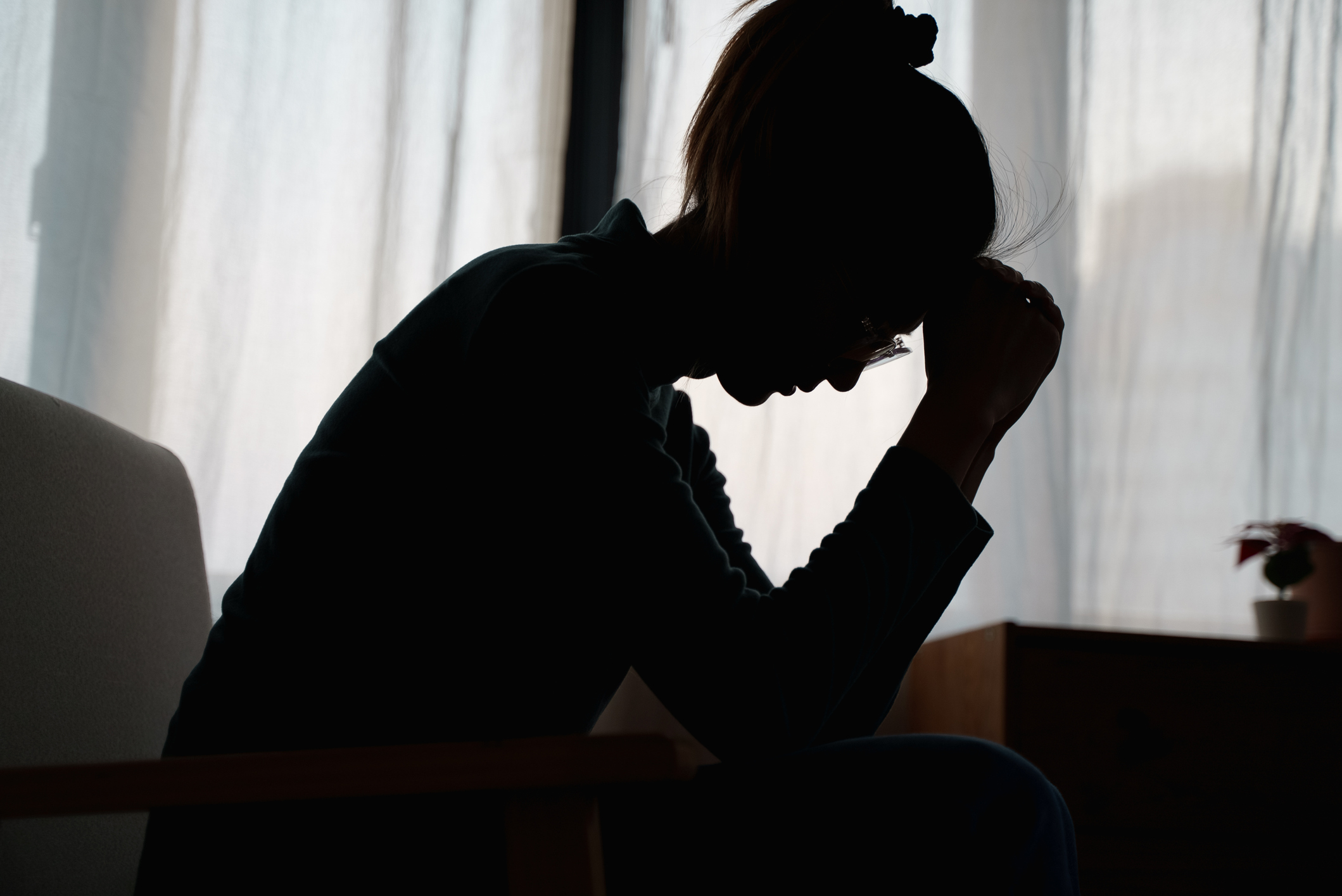 Silhouette photo of young Asian woman sitting alone in bedroom with her head bowed towards her closed hand