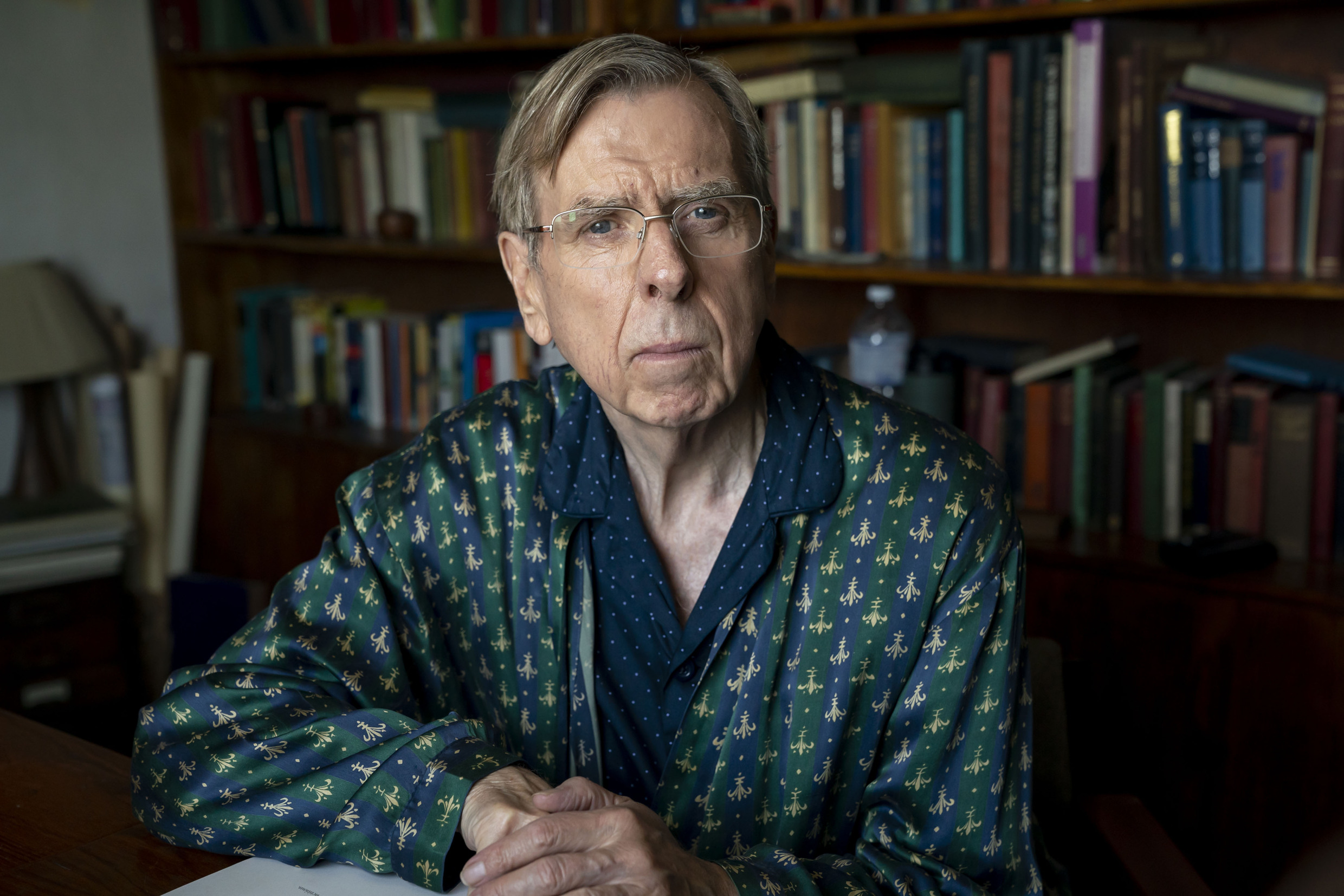 Timothy Spall in 'The Sixth Commandment' sitting in front of a book shelf looking suspicious